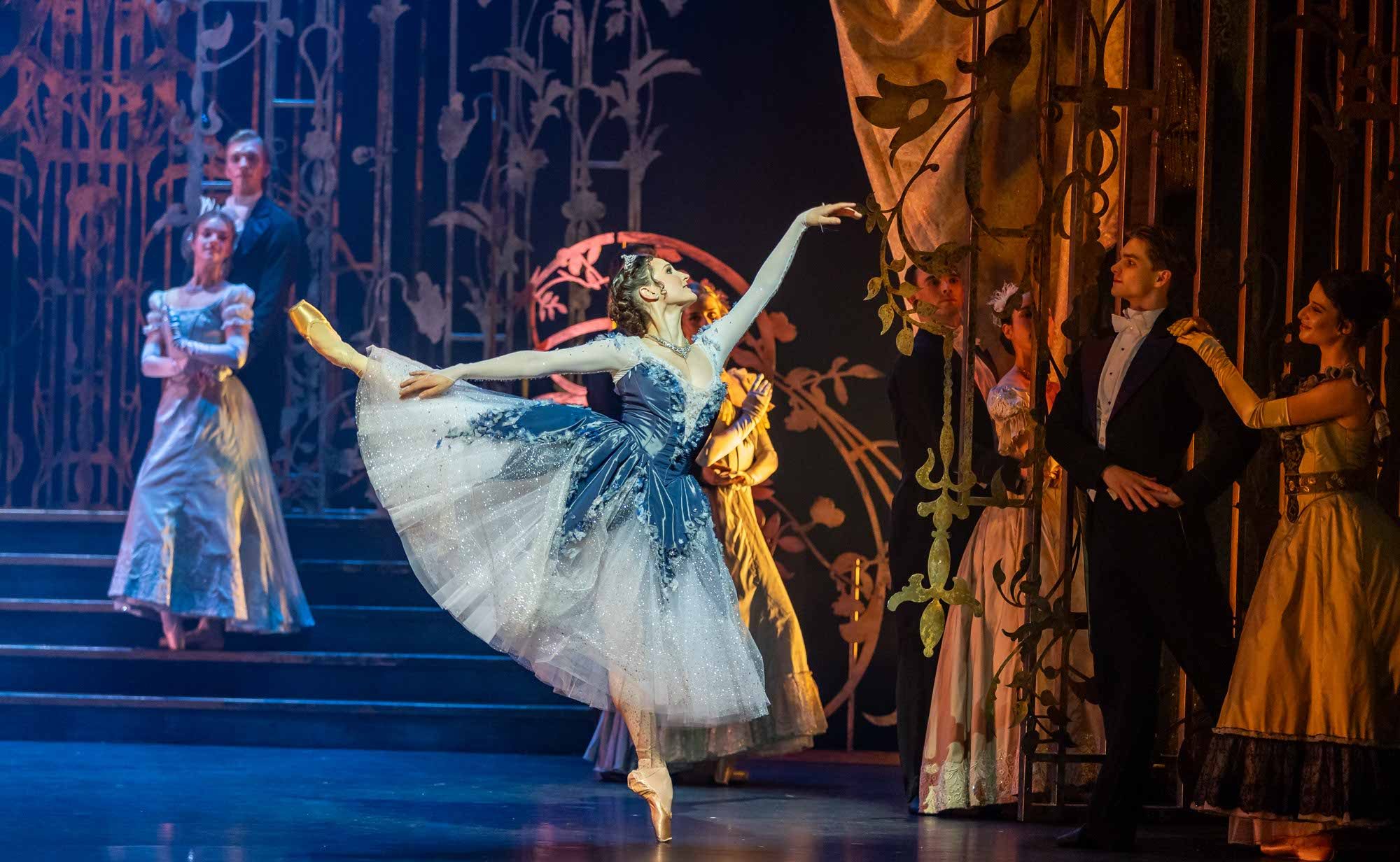 A ballerina in a blue and white dress performs an elegant arabesque on stage, surrounded by dancers in formal period costumes, with ornate golden set decorations in the background.